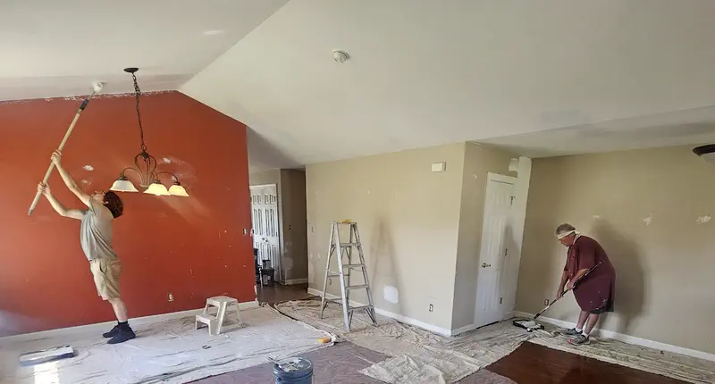 Two of the crew painting a vaulted living room, one rolling the ceiling by a terracotta wall, the other cutting in a wall, drop cloths down